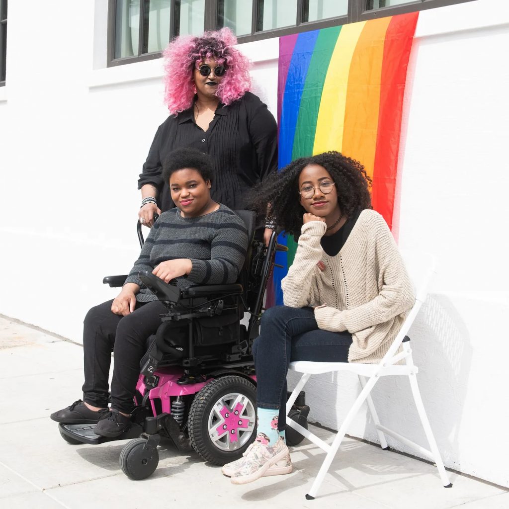 Three Black and disabled folx (a non-binary person holding a cane, a woman sitting in a power wheelchair, and a woman sitting in a chair) partially smiling at the camera while a rainbow pride flag drapes on the wall behind them.